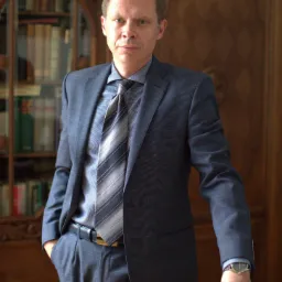 Portret mężczyzny w garniturze opierającego się o biurko z książką, na tle drewnianej biblioteczki.