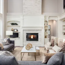 Salon z wysokim sufitem, kominkiem obudowanym białą cegłą i wkładem kominkowym z widocznym płomieniem, otoczony zabudowanymi półkami i dwiema sofami.