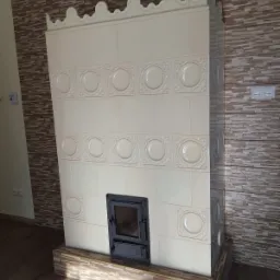 Biały, kaflowy piec z czarnymi drzwiczkami, ozdobiony wypukłymi, okrągłymi wzorami, ustawiony na podstawie z płytek imitujących drewno, z tłem ściany wyłożonej płytkami w odcieniach brązu.