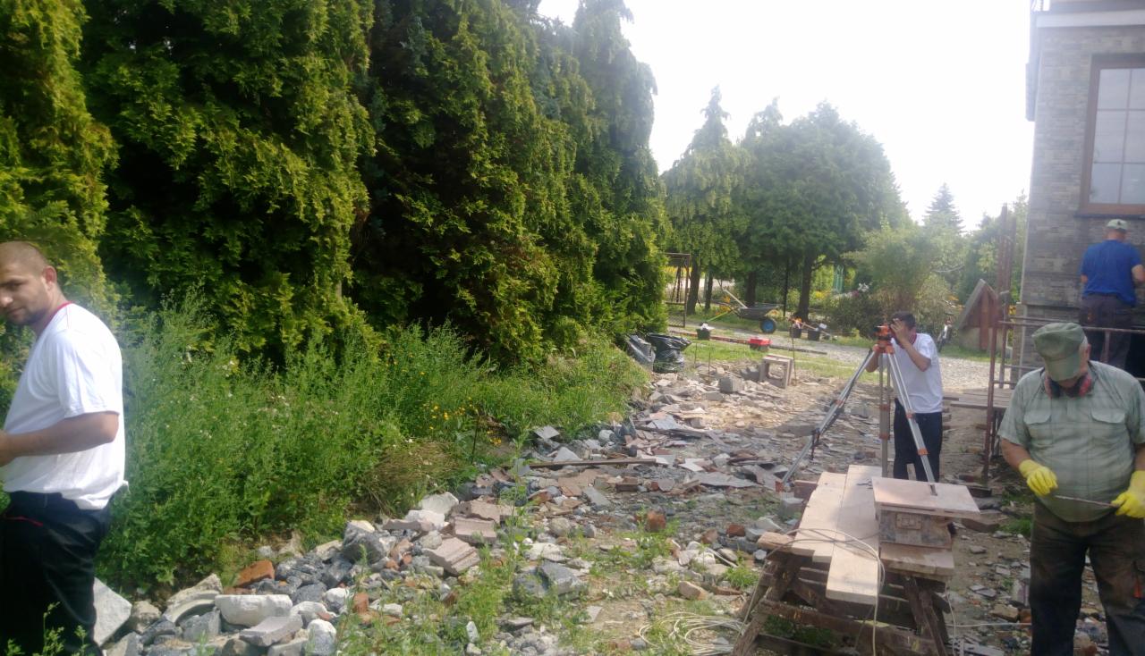 Prace przygotowawcze na działce: niwelacja terenu z użyciem niwelatora optycznego, widoczne gruzowisko, zieleń i pracownicy w trakcie pomiarów.