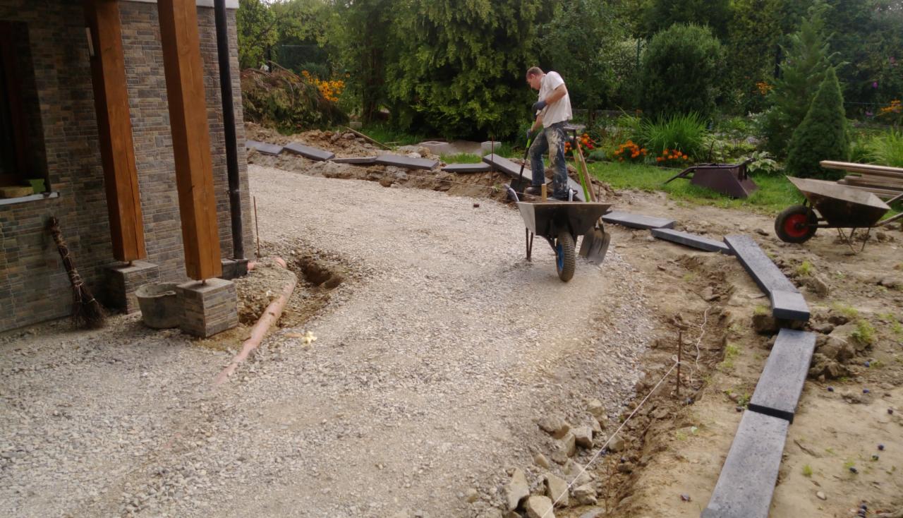 Pracownik w wózku transportuje materiał na podsypkę pod obrzeża z ciemnej kostki granitowej, widoczne wytyczone sznurkiem krawędzie przyszłego chodnika, w tle budynek z kamienną elewacją i zielenią.