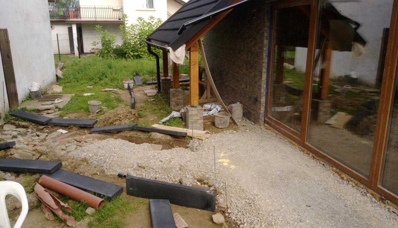Przygotowanie podłoża pod taras z kostki: widoczne wykopane fundamenty, podsypka żwirowa, ułożone obrzeża z ciemnej kostki, rury instalacyjne i narzędzia na trawie.