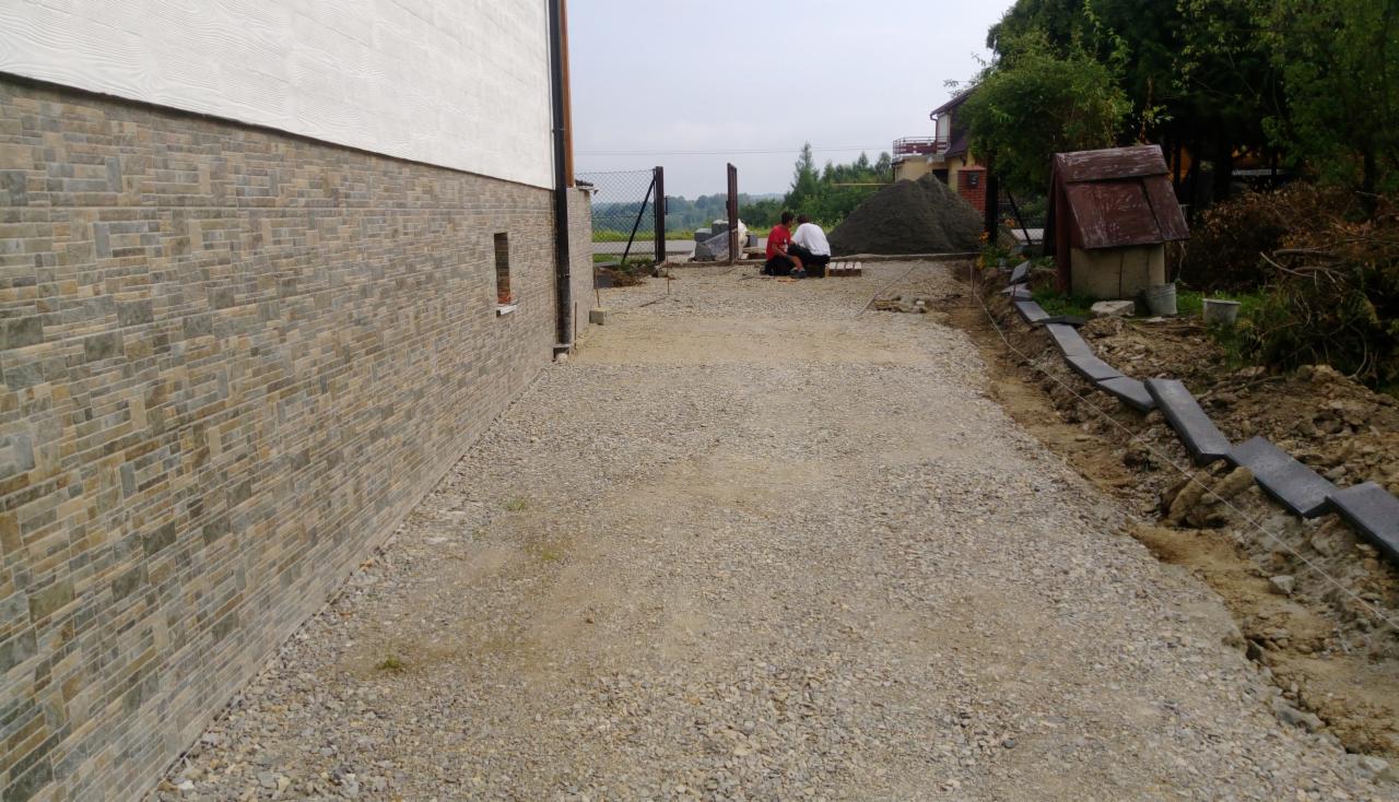 Przygotowanie podłoża pod kostkę brukową: wyrównany teren z podsypką, widoczni dwaj pracownicy przy ogrodzeniu, ułożone krawężniki z boku, dom z elewacją imitującą kamień w tle.