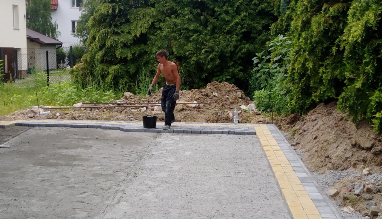 Mężczyzna bez koszuli w roboczych spodniach i rękawicach, z wiadrem, układa obrzeże z kostki brukowej w kolorze szarym i żółtym, na przygotowanym podłożu, w tle hałda ziemi i butelka wody.