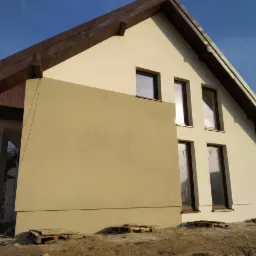 Dom jednorodzinny z częściowo wykończoną elewacją w dwóch odcieniach beżu, widoczne palety na ziemi, rusztowanie i fragment nieotynkowanej ściany.