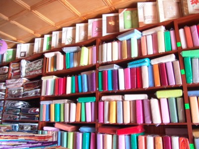 Półki wypełnione kolorowymi rolkami tkanin i kompletami pościeli w opakowaniach, widoczne w warsztacie krawieckim lub sklepie z tekstyliami.