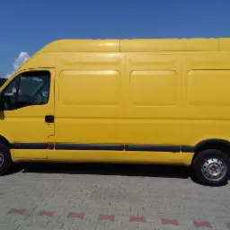Ż&oacute;łty samoch&oacute;d dostawczy Renault Master sfotografowany z boku, zaparkowany na szarej kostce brukowej z czerwonymi akcentami, na tle błękitnego nieba.