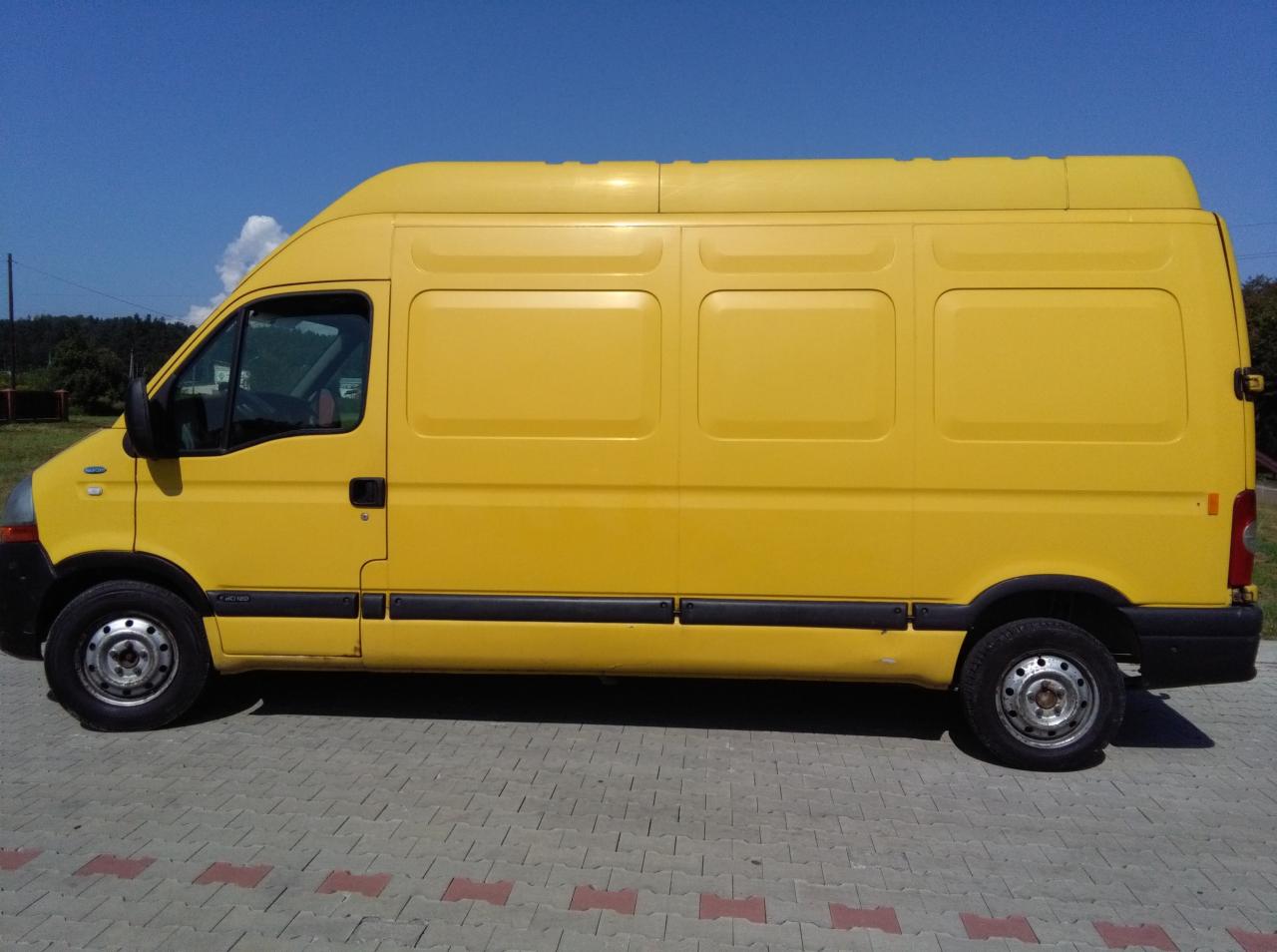 Żółty samochód dostawczy Renault Master sfotografowany z boku, zaparkowany na szarej kostce brukowej z czerwonymi akcentami, na tle błękitnego nieba.