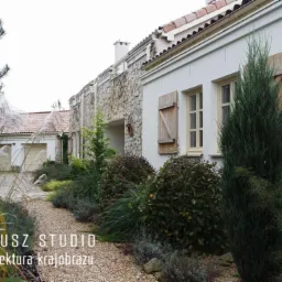 Ogród w stylu toskańskim z białym budynkiem, kamienną elewacją, drewnianymi okiennicami i ścieżką z drobnego żwiru, ozdobiony trawami ozdobnymi i niskimi krzewami.