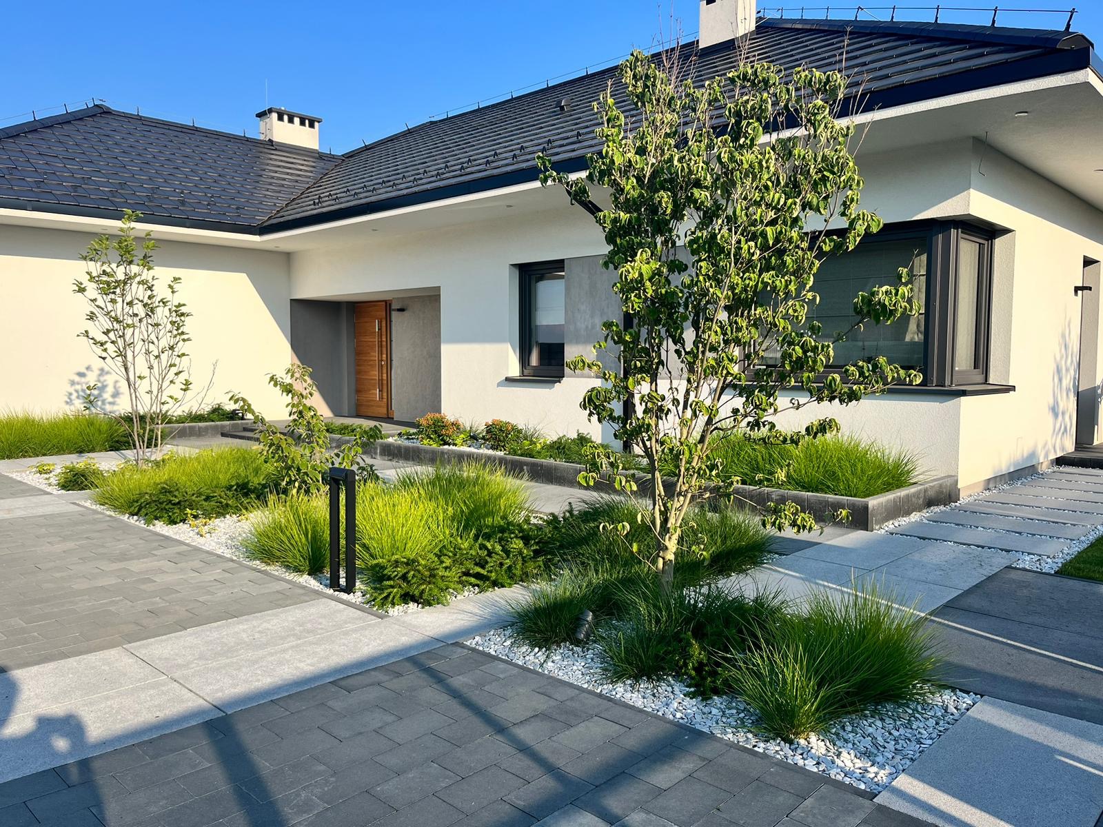Nowoczesny ogród z geometrycznymi rabatami, ozdobnymi trawami i ścieżką z kostki brukowej prowadzącą do domu z ciemną dachówką w słoneczny dzień.