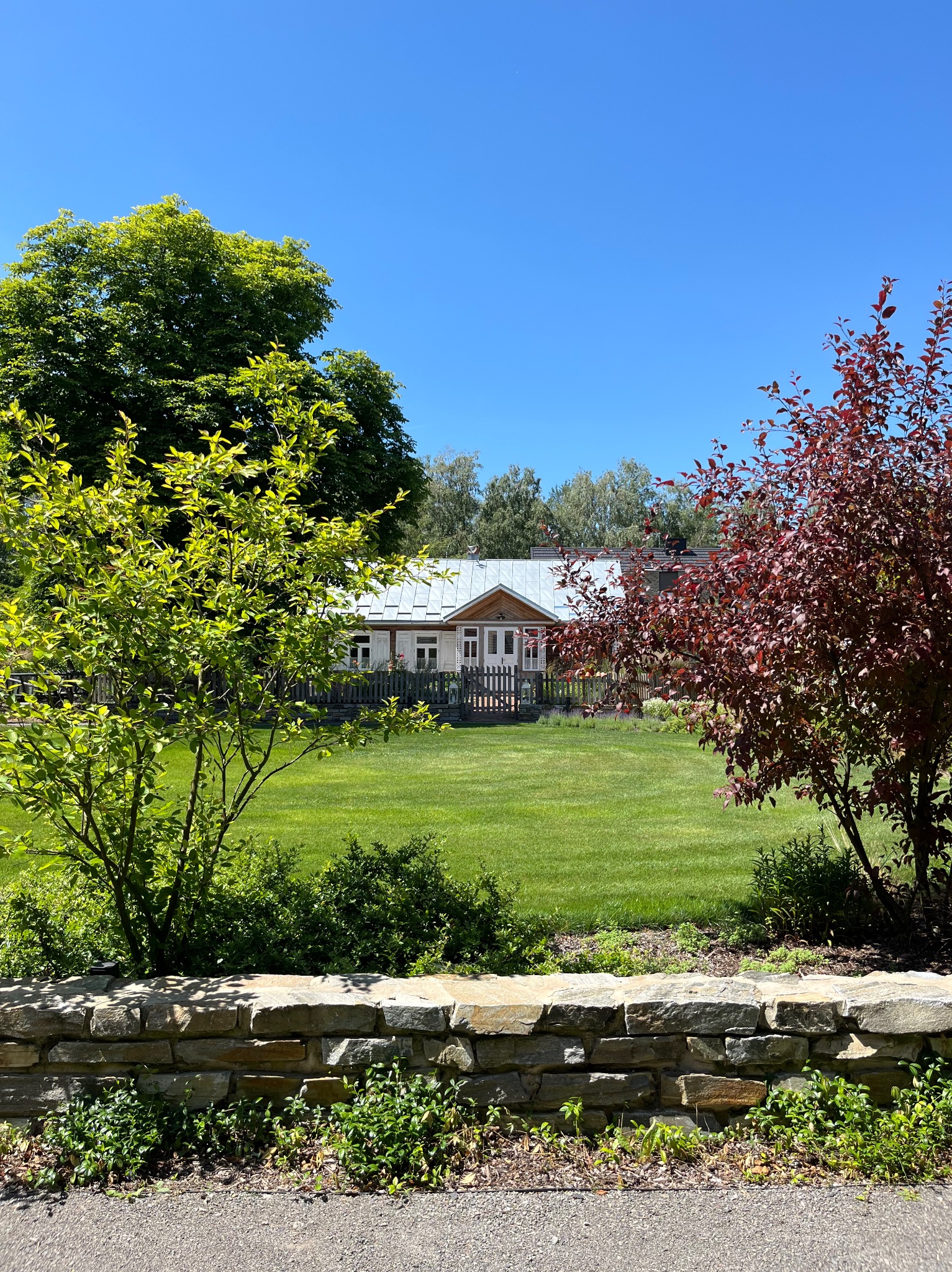Zadbany trawnik przed domem z szarym dachem, otoczony zielenią i drzewami o czerwonych liściach. Kamienny murek w pierwszym planie. Słoneczny dzień, błękitne niebo.