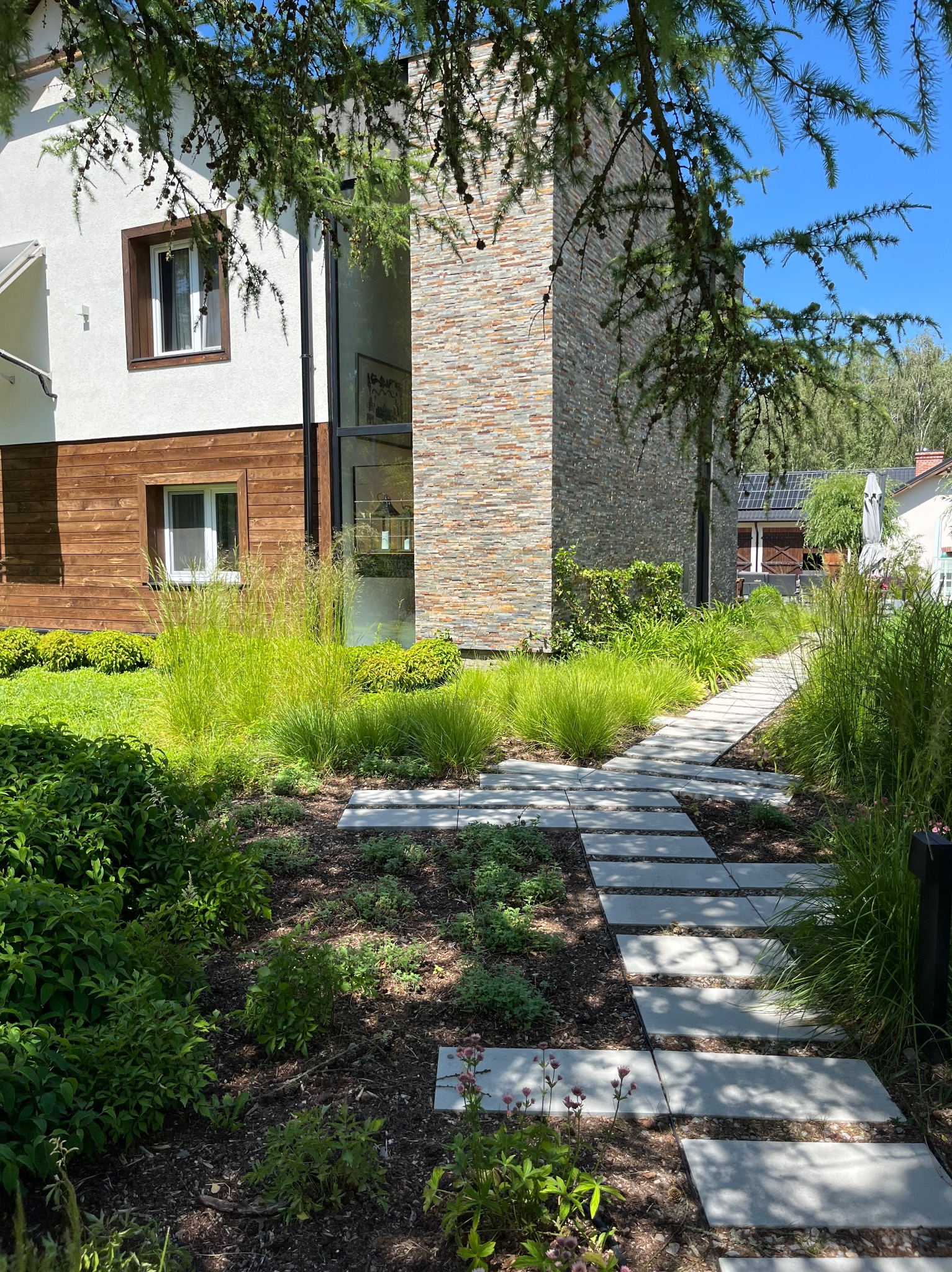 Nowoczesny ogród z ścieżką z płyt betonowych prowadzącą do domu z elewacją z kamienia i drewna. Zadbana zieleń, trawy ozdobne i krzewy.