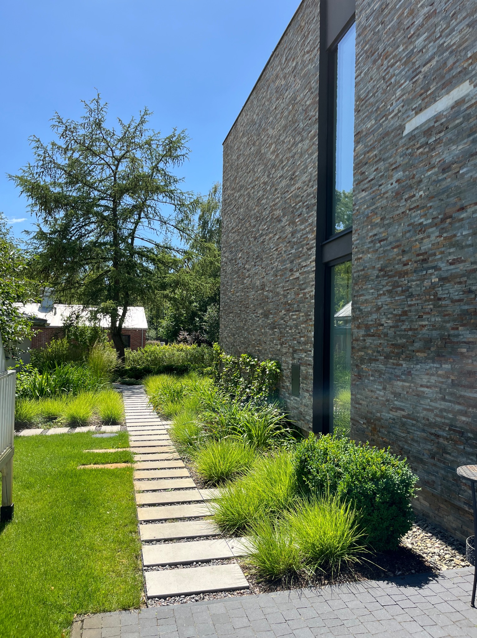 Nowoczesny ogród z kamienną ścieżką i ozdobnymi trawami prowadzącą wzdłuż elewacji budynku wykończonej kamieniem. Minimalistyczny design z elementami zieleni.