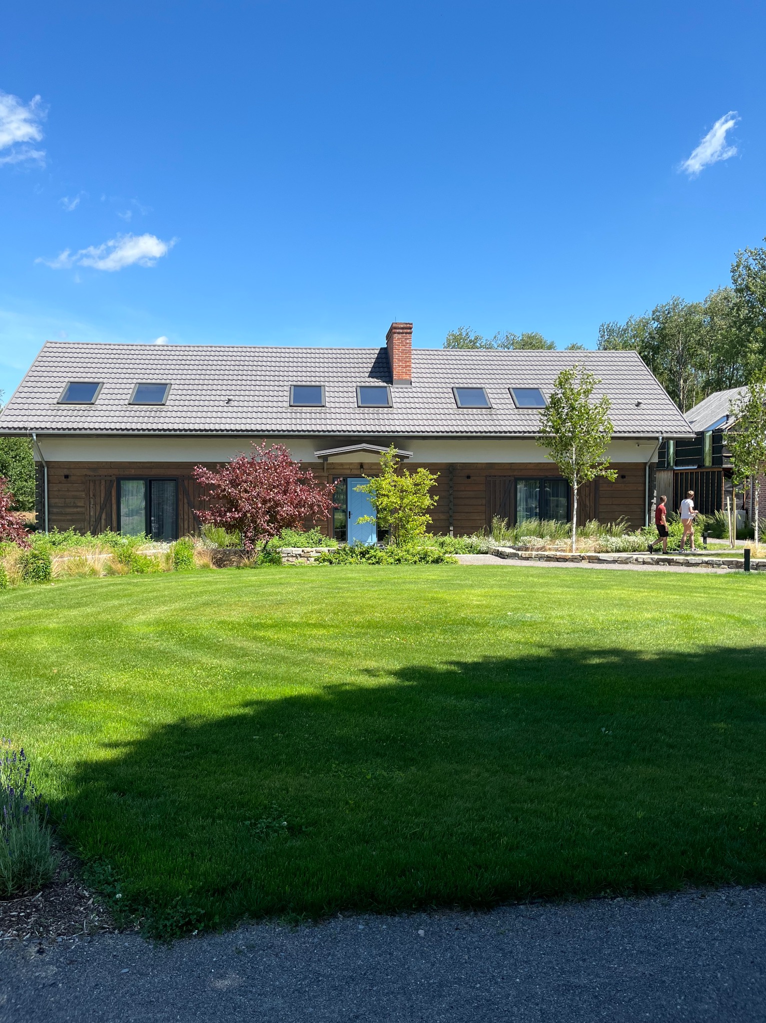 Rozległy trawnik przed domem z drewnianą elewacją i niebieskimi drzwiami. Uwagę przykuwają starannie dobrane rośliny i drzewa, tworzące harmonijną kompozycję ogrodu.