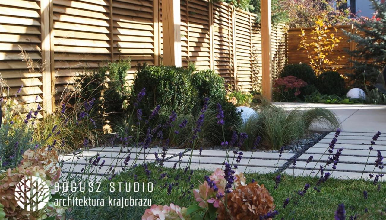 Nowoczesny ogród z drewnianym ażurowym ogrodzeniem, geometrycznymi ścieżkami z betonowych płyt i nasadzeniami z lawendy, bukszpanu i traw ozdobnych; w tle widoczne kule dekoracyjne.