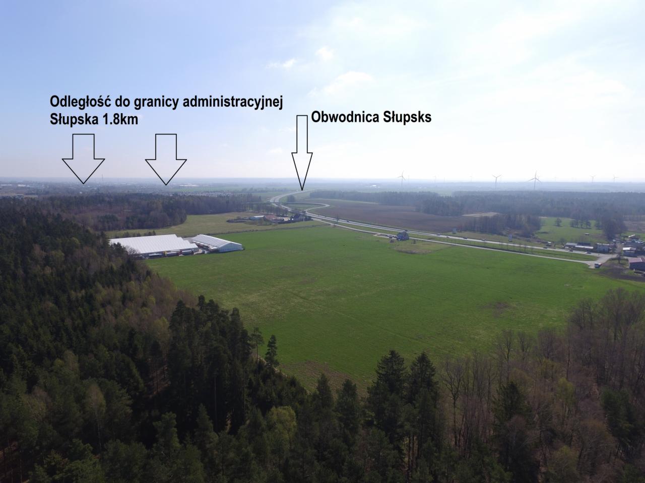 Szeroki widok z lotu ptaka na teren w pobliżu Słupska, z widocznymi polami, lasami, obwodnicą i budynkami magazynowymi; w tle turbiny wiatrowe.