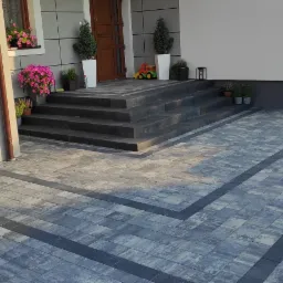 Nowoczesna kostka brukowa w odcieniach szarości z ciemnymi pasami, prowadząca do schodów wejściowych domu z kwiatami i dekoracjami.