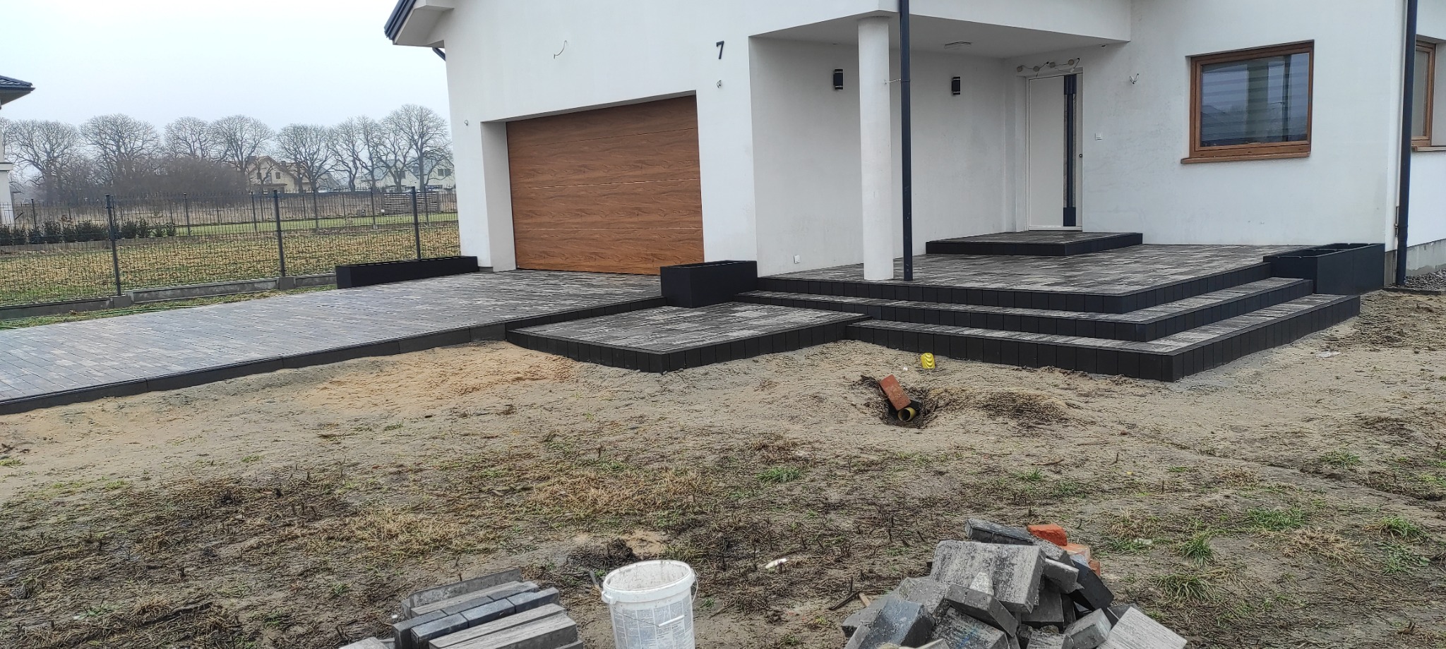 Nowoczesny podjazd i schody wejściowe z grafitowej kostki brukowej przy białym domu z brązową bramą garażową. Widoczne niedokończone prace ziemne.