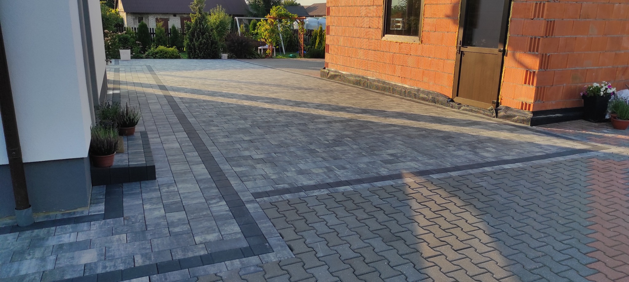 Ułożona kostka brukowa w odcieniach szarości z ciemniejszymi obramowaniami. W tle budynek w stanie surowym otwartym i zieleń ogrodu.