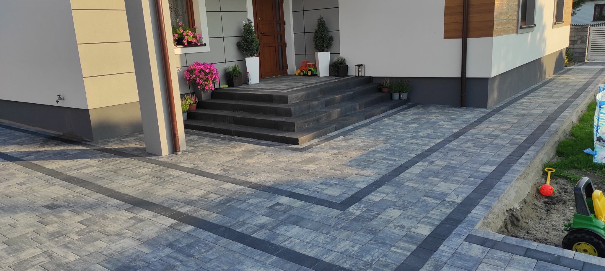 Nowoczesna kostka brukowa w odcieniach szarości z ciemnymi pasami, prowadząca do schodów wejściowych domu z kwiatami i dekoracjami.
