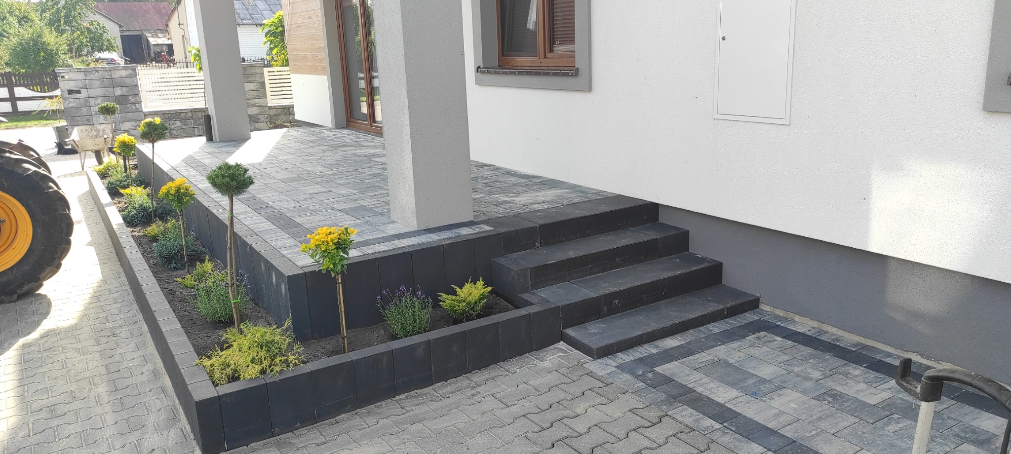 Nowoczesny podjazd z kostki brukowej w odcieniach szarości, schody z ciemnych kostek, rabata z roślinami i fragment traktora z lewej strony kadru.