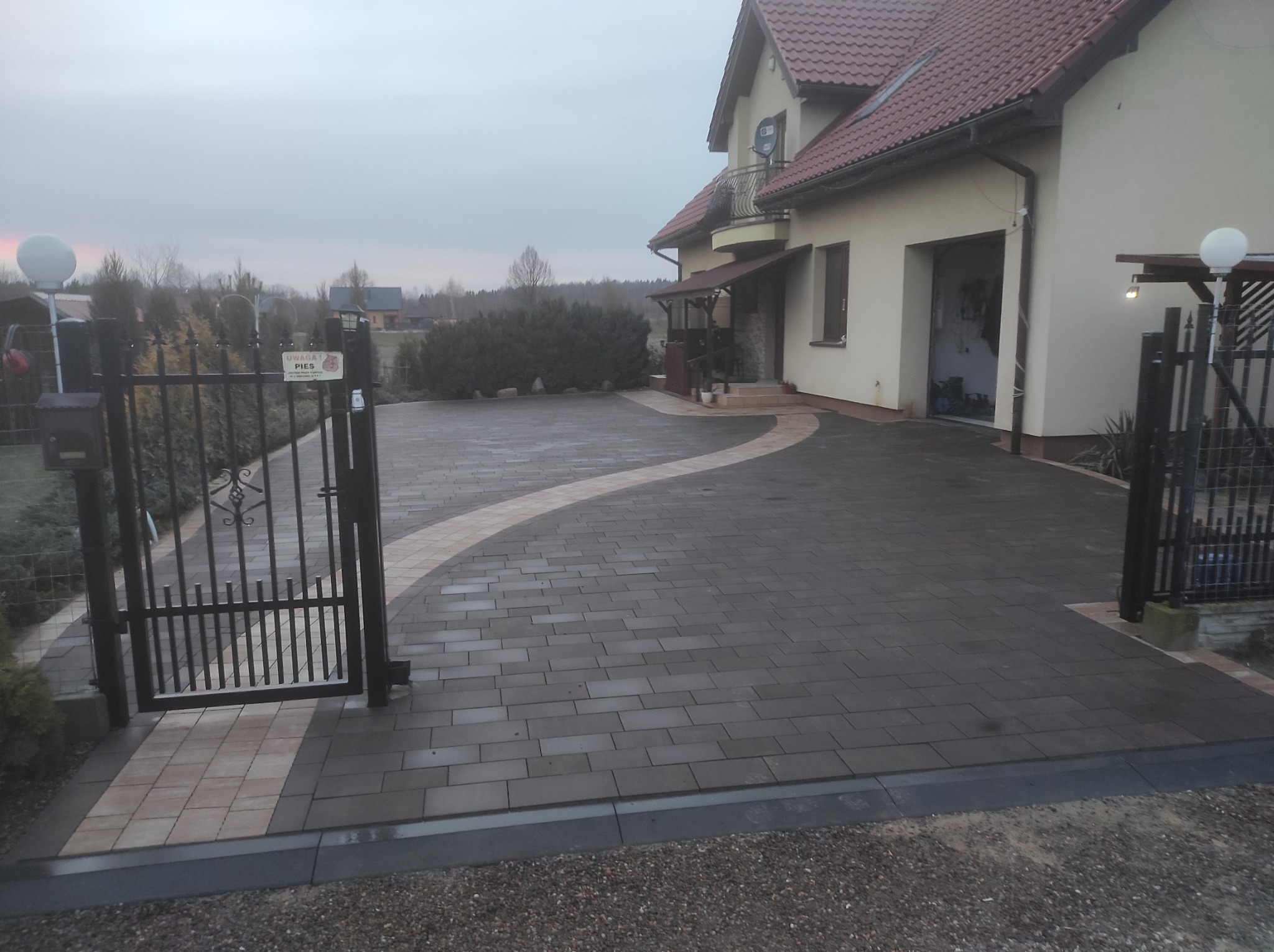 Ułożona, ciemna kostka brukowa przed domem jednorodzinnym z elementami dekoracyjnymi w postaci jasnej kostki. Widoczna brama wjazdowa i zadbany ogród.