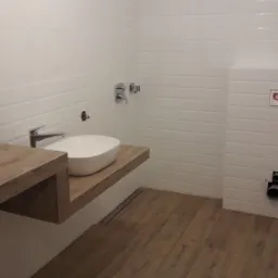 Nowoczesna łazienka z białymi, prostokątnymi płytkami na ścianach, drewnianą podłogą i blatem pod umywalkę, chromowana bateria umywalkowa, oraz widoczne elementy instalacji sanitarnej