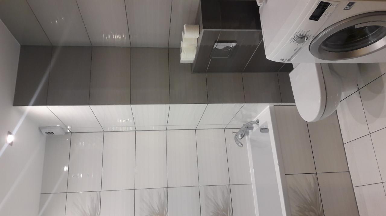 Nowoczesna łazienka z szarymi i białymi płytkami, wiszącą toaletą, pralką Electrolux i ukrytym oświetleniem punktowym.