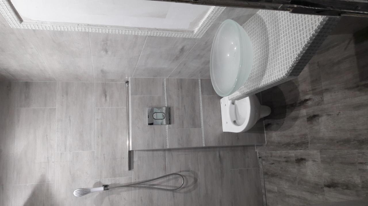 Nowoczesna toaleta z misą umywalkową na blacie z mozaiki, szare płytki imitujące drewno na ścianach i podłodze, deszczownica z chromowaną armaturą.