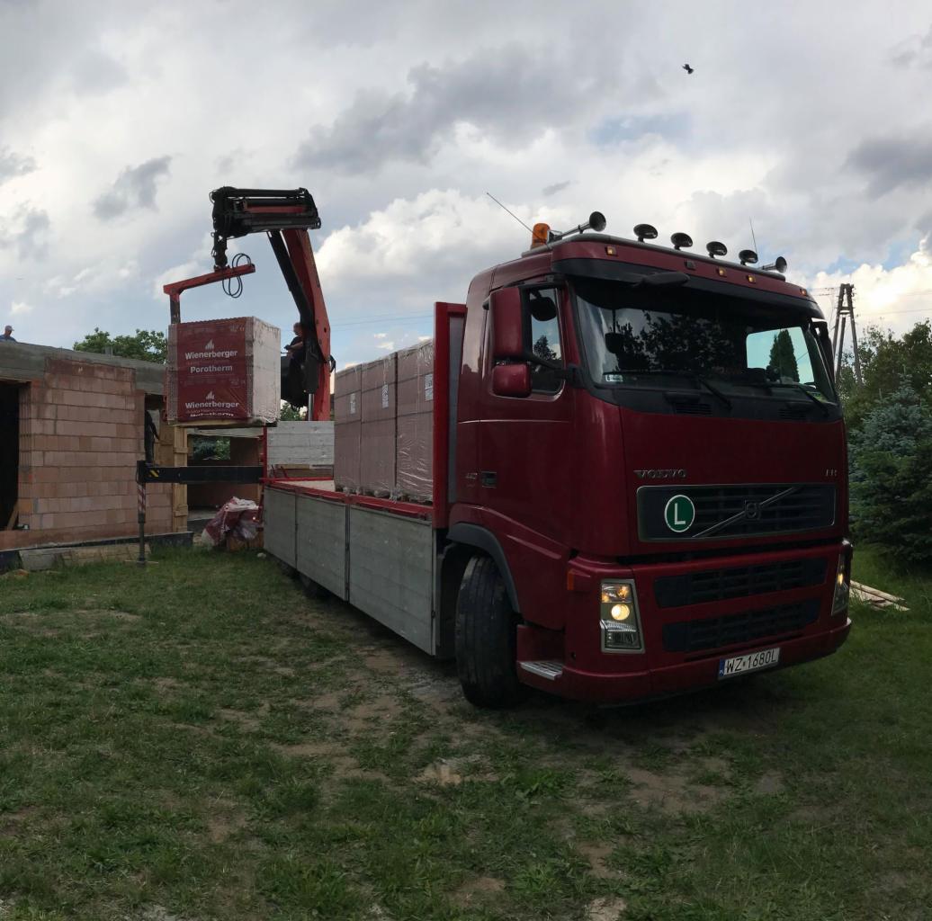Czerwona ciężarówka Volvo z dźwigiem rozładowuje palety z cegłami Wienerberger Porotherm na placu budowy domu z czerwonej cegły.