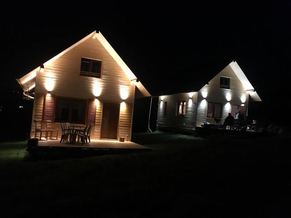 Dwa oświetlone domki z drewna w nocy, widoczne stoły i krzesła na tarasach, ciemne tło.