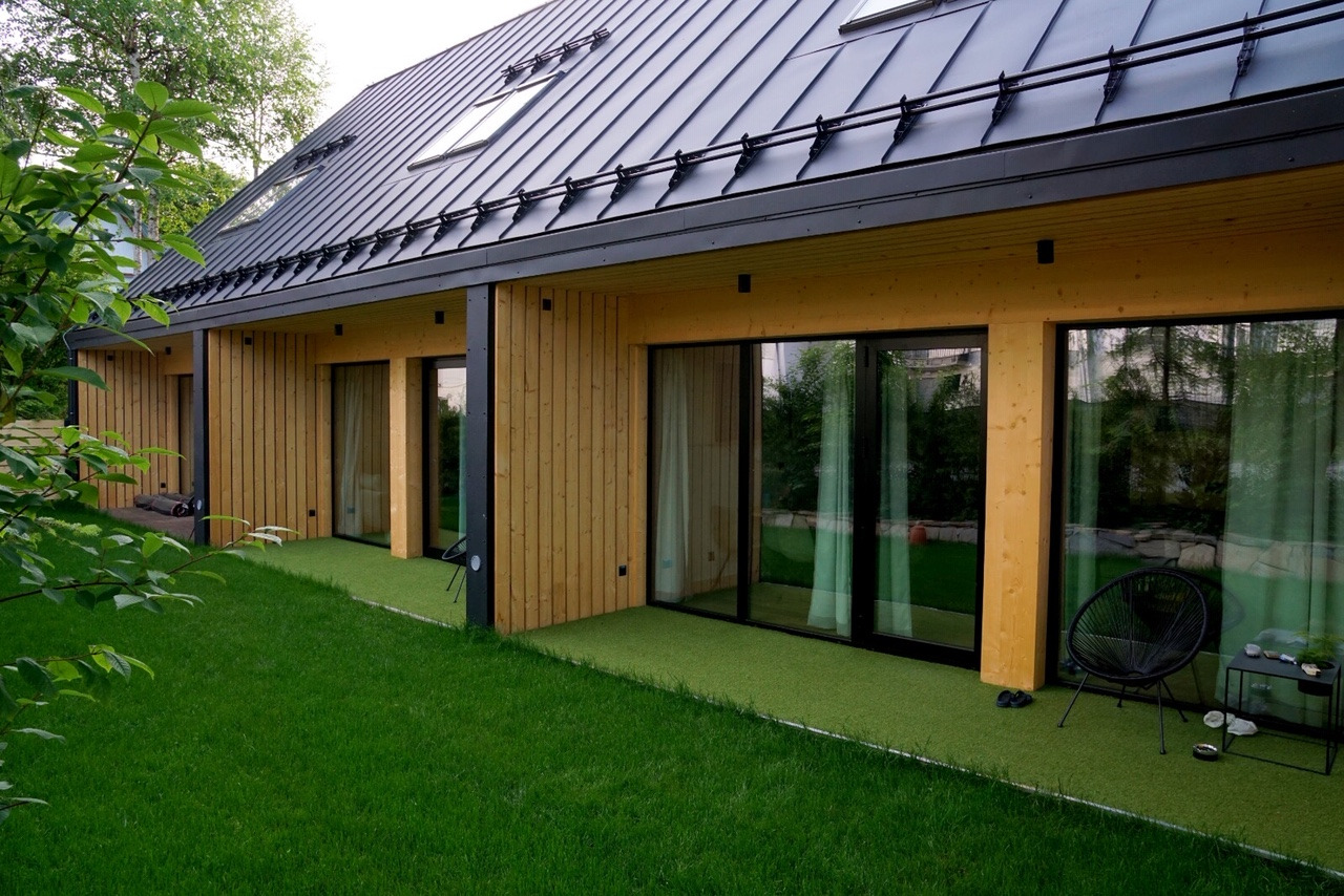 Nowoczesny dom szkieletowy z czarnym dachem na rąbek stojący, drewnianą elewacją i dużymi oknami przesuwnymi wychodzącymi na trawnik.