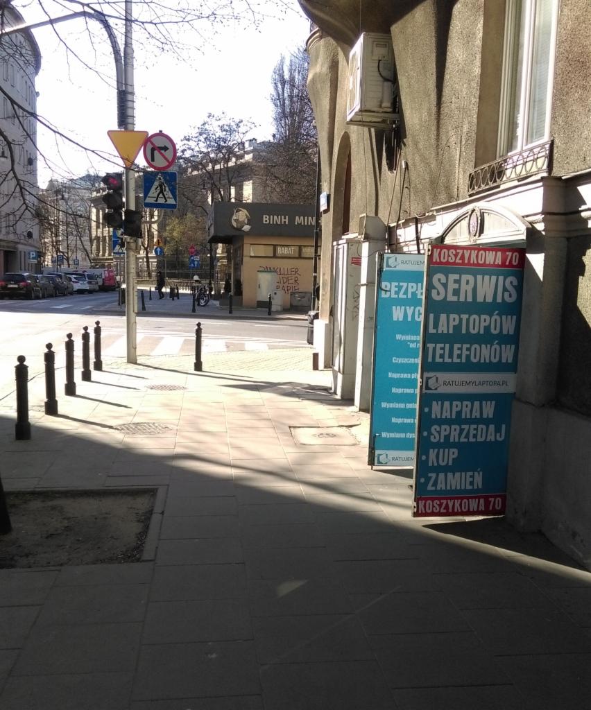 Reklama serwisu laptopów i telefonów na tablicach wystawionych przed budynkiem przy ulicy Koszykowej 70, widok na chodnik i ulicę w słoneczny dzień.