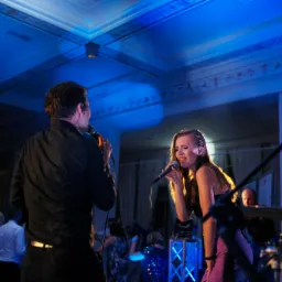 Wokalistka z mikrofonem śpiewa na scenie oświetlonej niebieskim światłem, obok wokalista, w tle widoczni goście.