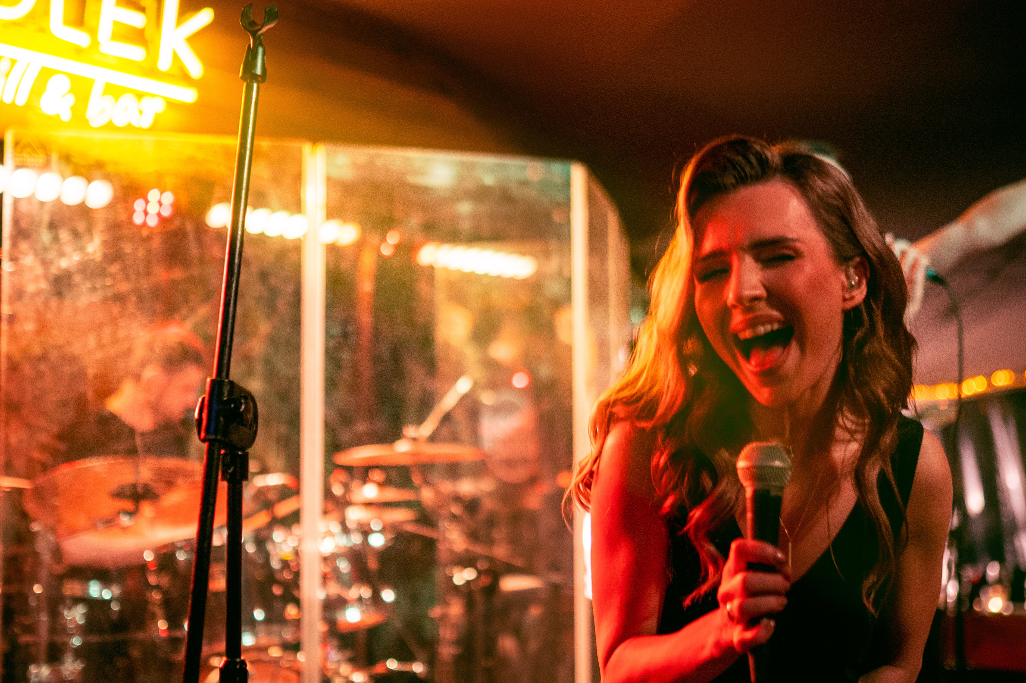 Wokalistka z długimi włosami śpiewa do mikrofonu na tle rozmytej perkusji i neonowego napisu 'LEK grill & bar' w ciepłym, czerwonym oświetleniu.