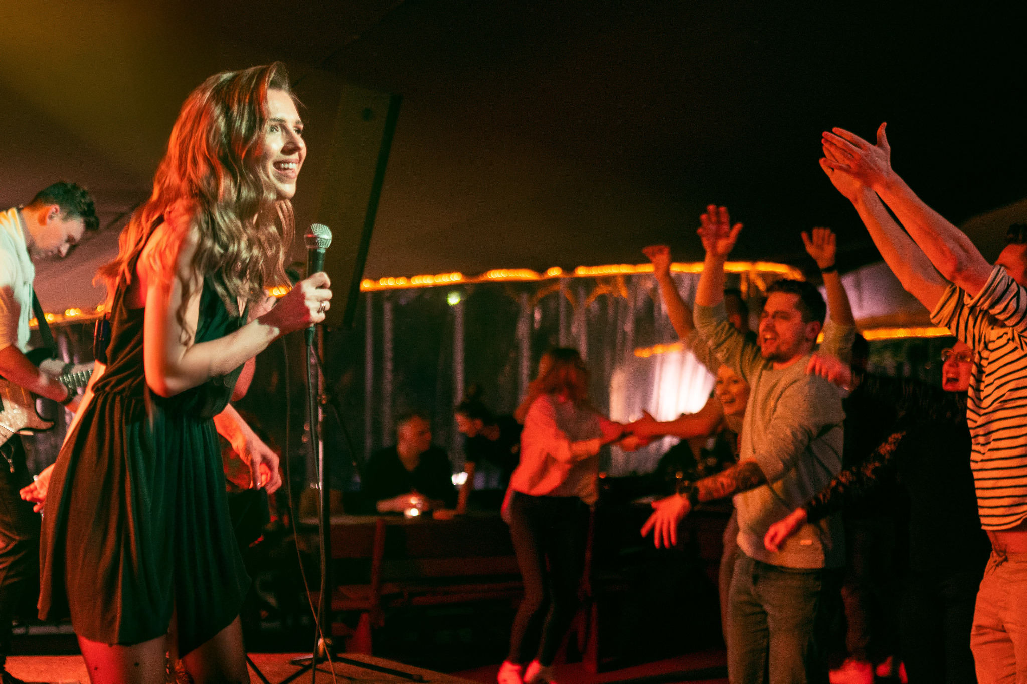 Wokalistka w zielonej sukience śpiewa do mikrofonu na scenie, w tle gitarzysta i rozbawiony tłum z podniesionymi rękami.