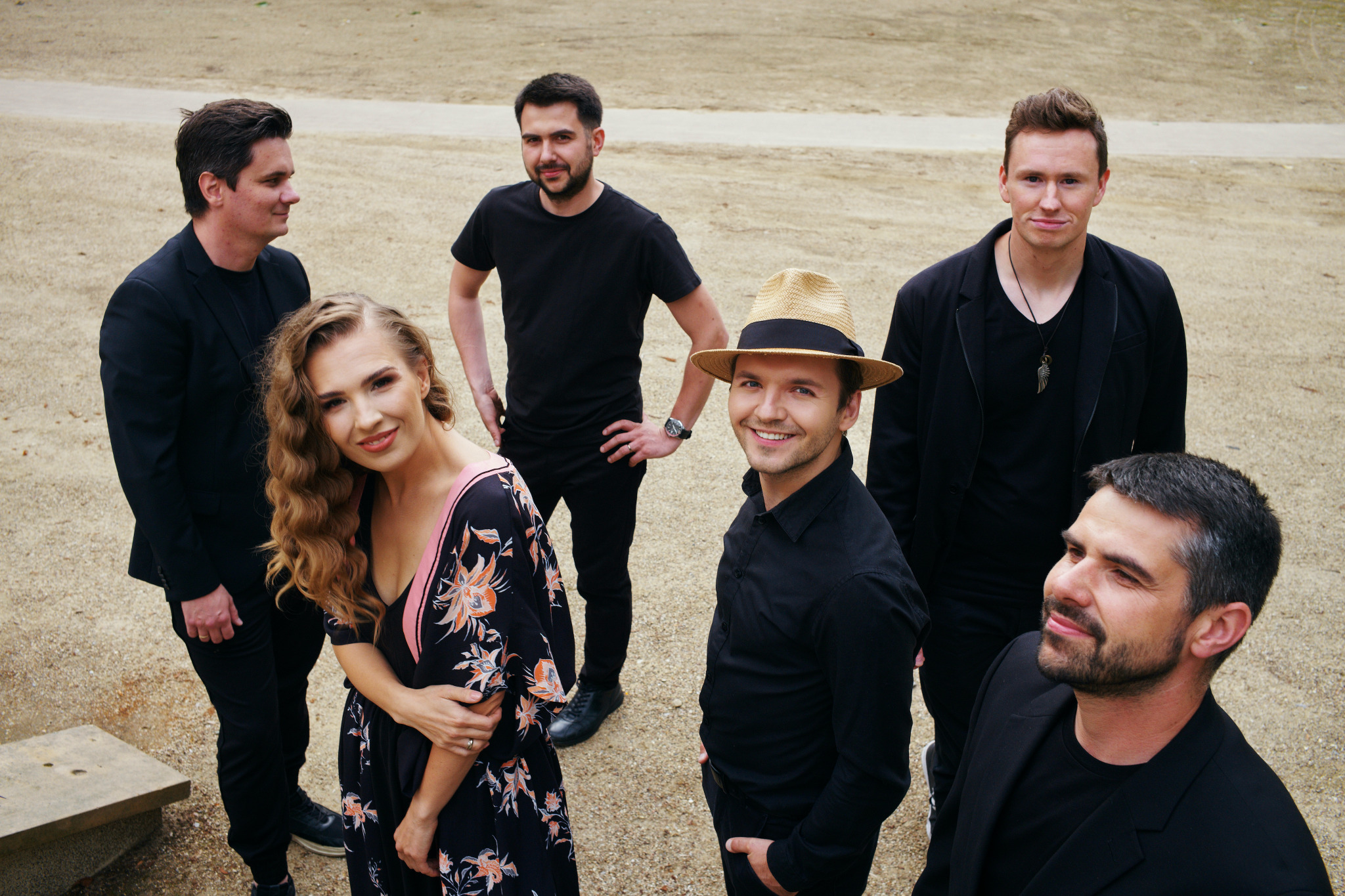 Zespół muzyczny w czarnych strojach i słomkowym kapeluszu pozuje do zdjęcia na tle piaszczystego placu.