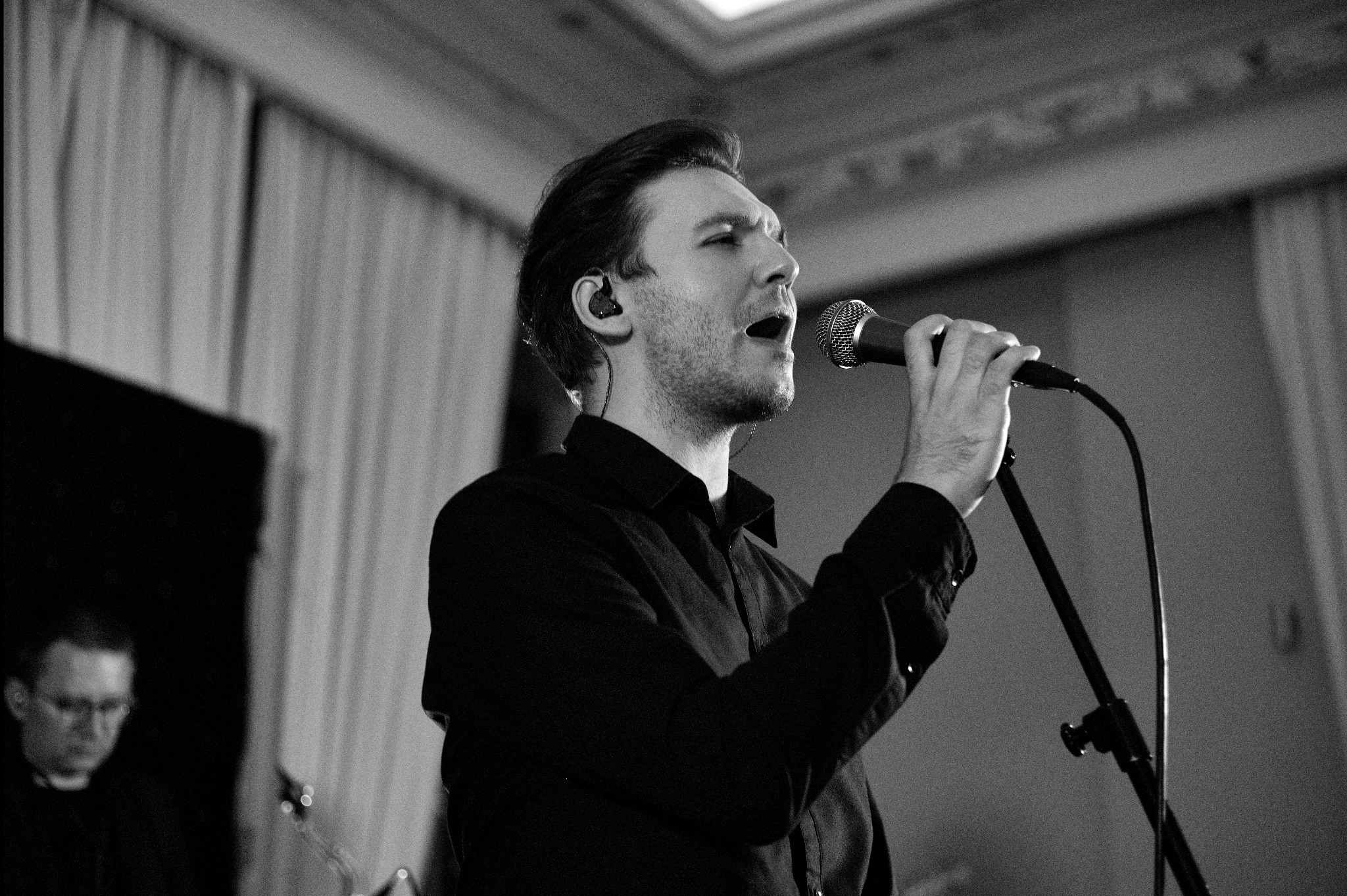 Czarno-białe zdjęcie wokalisty z mikrofonem na statywie, z wkładką douszną, podczas występu na scenie z zasłonami w tle i zarysami innych muzyków.