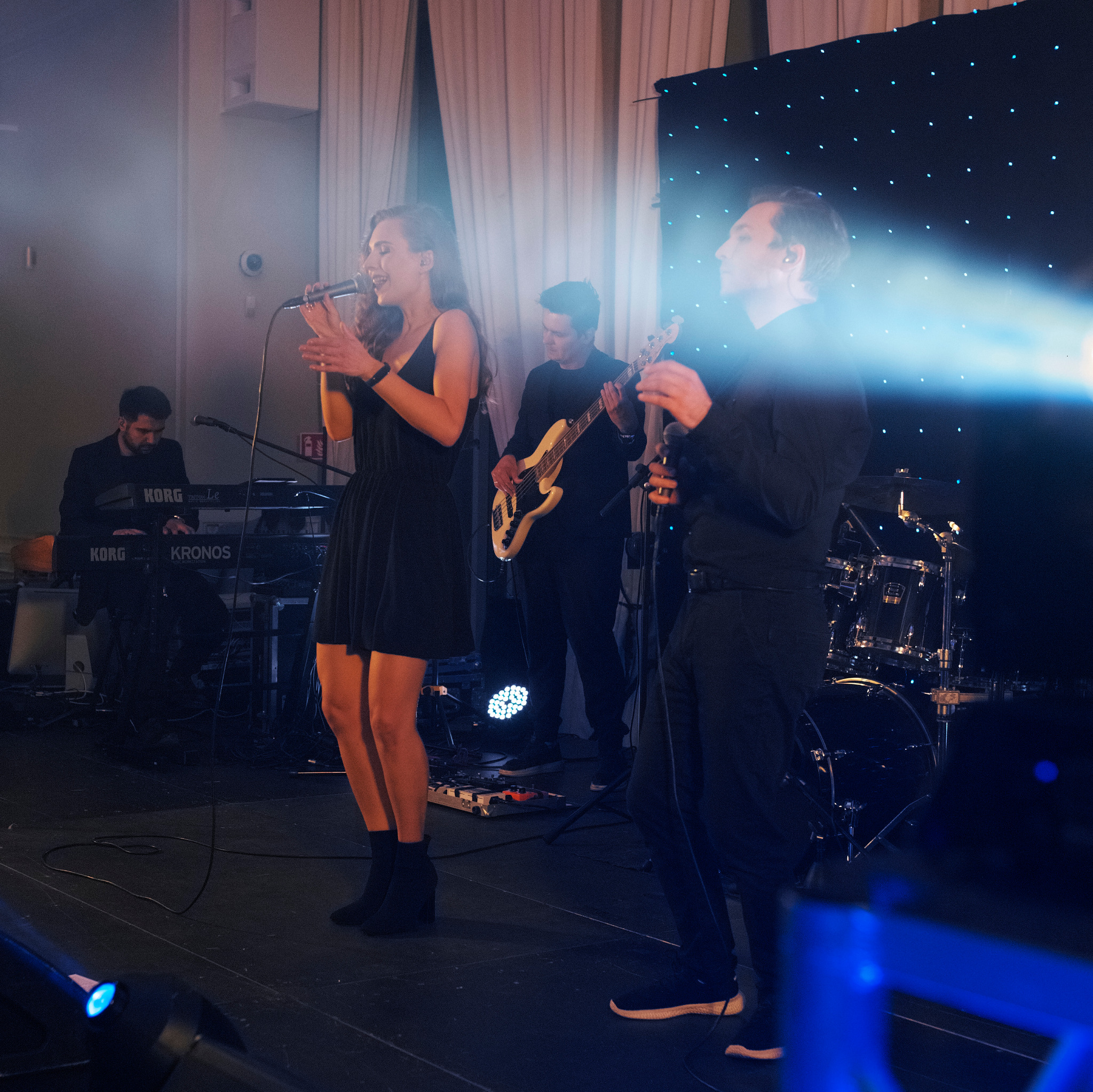 Występujący na scenie muzycy: wokalistka w czarnej sukience śpiewa do mikrofonu, gitarzysta gra na basie, klawiszowiec gra na Korg Kronos, za nimi dekoracja z kurtyny i światełek.