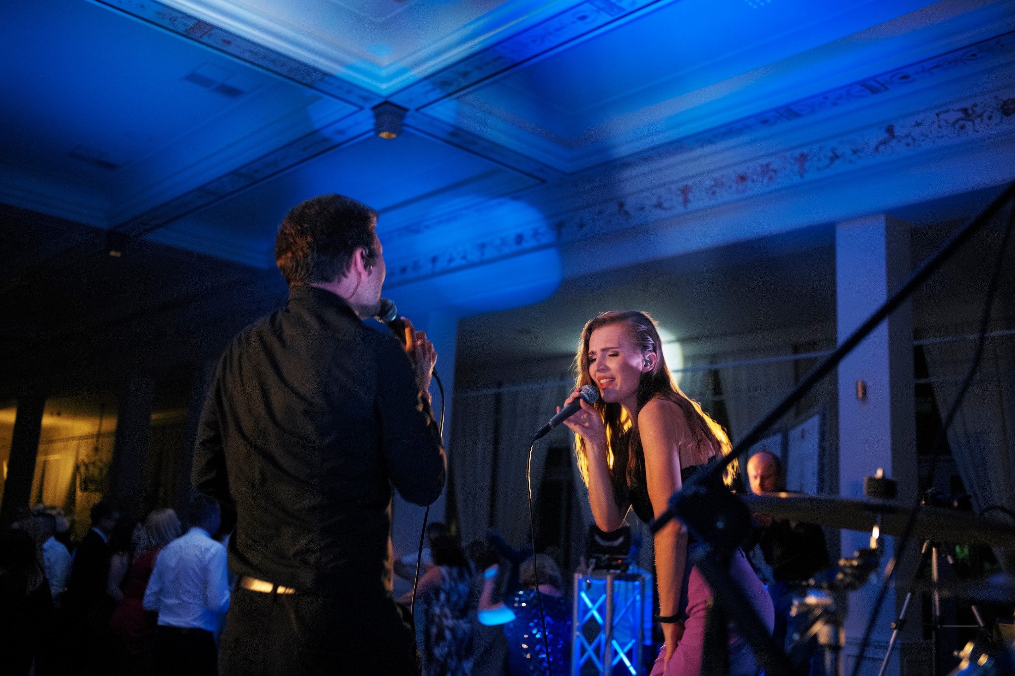 Wokalistka z mikrofonem śpiewa na scenie oświetlonej niebieskim światłem, obok wokalista, w tle widoczni goście.