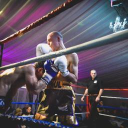 JMI Fotografia - Dwóch bokserów w ringu podczas walki, oświetlenie sceniczne w odcieniach fioletu i różu, w tle widoczna publiczność i sędzia.