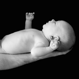 JMI Fotografia - Czarno-białe zdjęcie noworodka leżącego na przedramieniu dorosłego, z jedną rączką uniesioną w górę, na jednolitym czarnym tle.