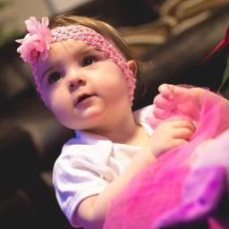 JMI Fotografia - Urocza dziewczynka z różową opaską i kwiatkiem, trzymająca w rączkach kawałek różowego tiulu, spogląda w górę.