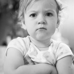 JMI Fotografia - Czarno-białe portretowe ujęcie rozzłoszczonej dziewczynki z założonymi rękami, lekko zmarszczone brwi, na pierwszym planie fragment pasiastych spodni.