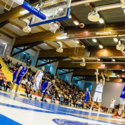JMI Fotografia - Mecz koszykówki: zawodnik w białym stroju oddaje rzut do kosza, w tle trybuny pełne widzów i oświetlenie hali.