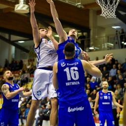 JMI Fotografia - Zawodnicy koszykówki w trakcie skoku do kosza, piłka w powietrzu, widownia w tle.