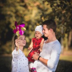 JMI Fotografia - Uśmiechnięta rodzina w parku: mama trzyma kolorowy wiatraczek, tata tuli roześmianą córeczkę w czerwonej sukience i białej czapeczce.