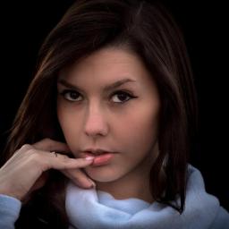 JMI Fotografia - Portret młodej kobiety z ciemnymi włosami, delikatny makijaż oczu, zamyślony wyraz twarzy, palec wskazujący na ustach, na czarnym tle.