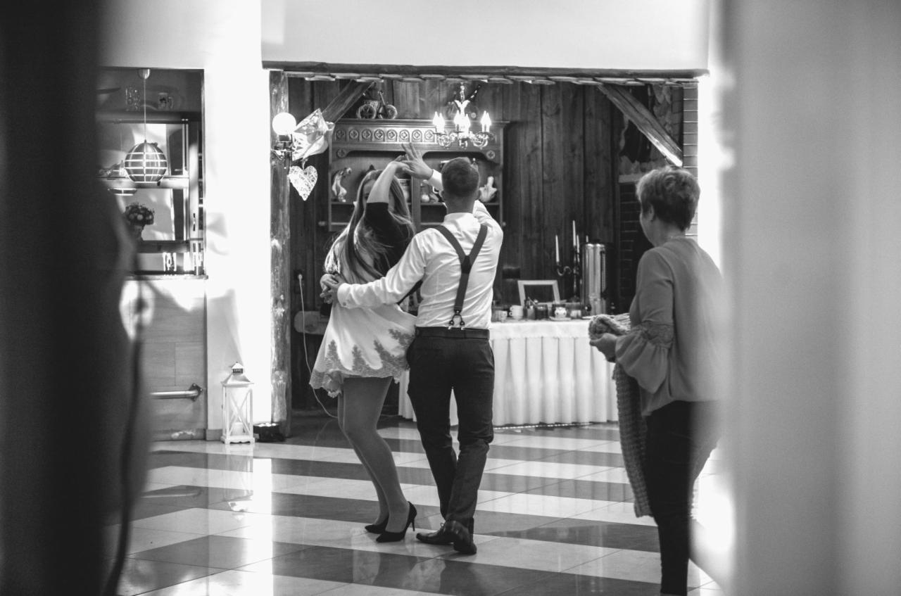 Czarno-białe zdjęcie pary tańczącej na parkiecie w paski, mężczyzna w szelkach, kobieta w krótkiej sukience, w tle bufet i drewniana ściana, z boku widoczna starsza kobieta.