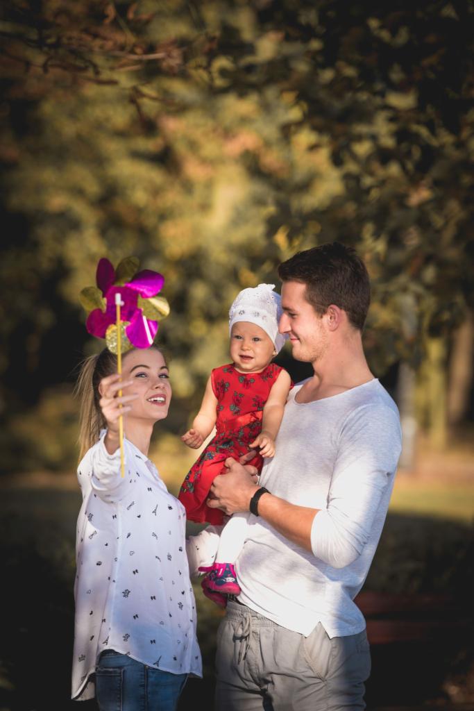 Uśmiechnięta rodzina w parku: mama trzyma kolorowy wiatraczek, tata tuli roześmianą córeczkę w czerwonej sukience i białej czapeczce.